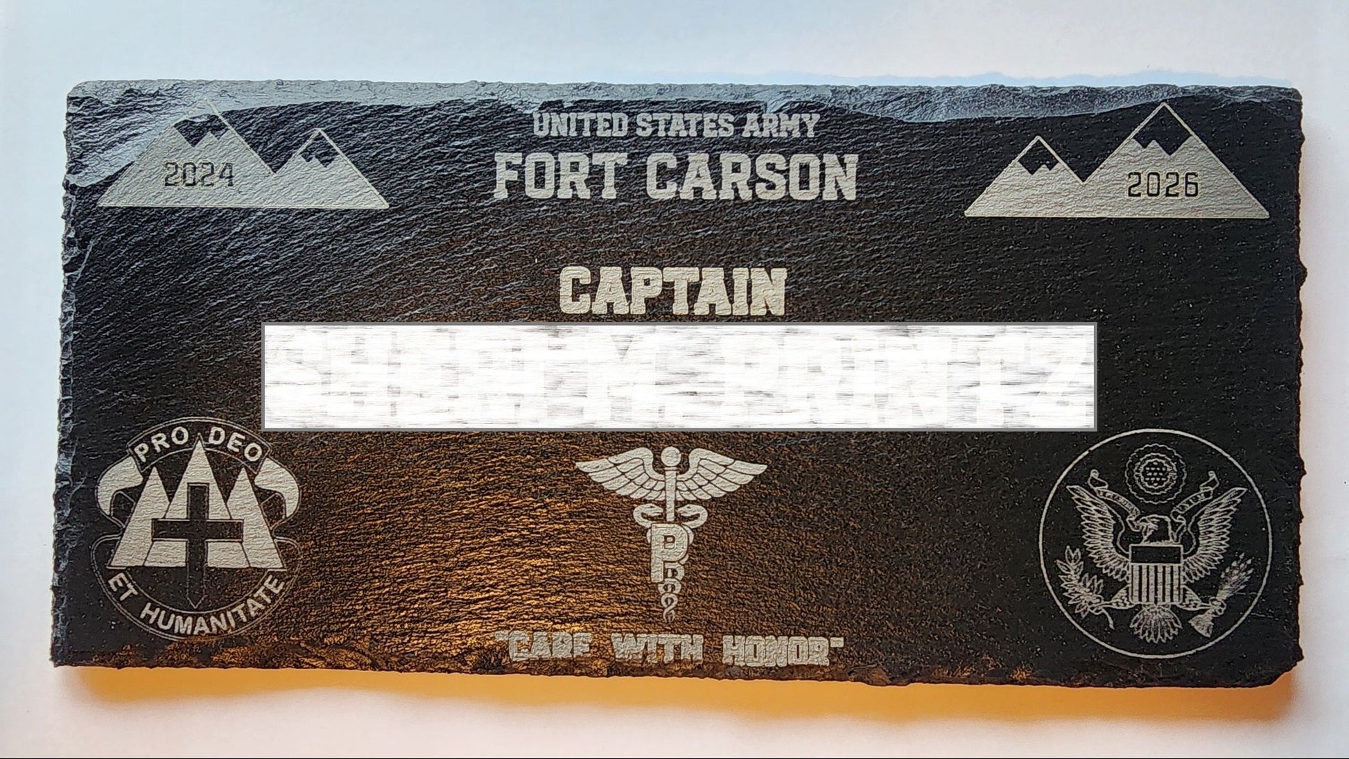 A black slate military commendation plaque for the United States Army Fort Carson, featuring engraved silver text, mountain graphics, and military insignia, with a white bar covering the central name area.