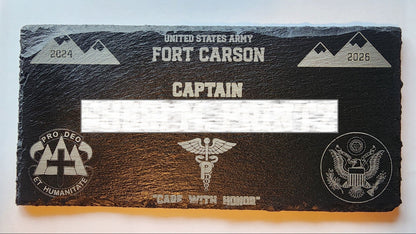 A black slate military commendation plaque for the United States Army Fort Carson, featuring engraved silver text, mountain graphics, and military insignia, with a white bar covering the central name area.