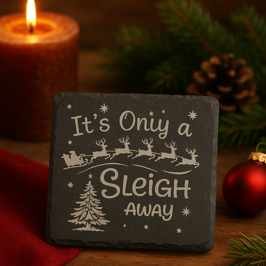 Slate Christmas coaster with 'It's Only a Sleigh Away' text, surrounded by festive decor on a wooden surface.